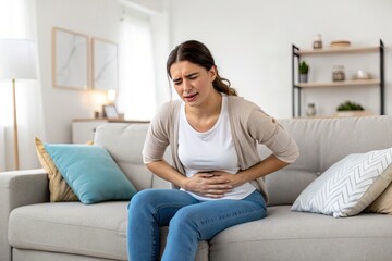Obraz premium Woman sitting on a couch in a bright living room, experiencing abdominal pain, looking distressed