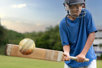 Young boy wearing a helmet plays cricket on a grassy field, focusing intensely as he hits the ball with a wooden bat. Outdoor sports and youth energy captured in motion.