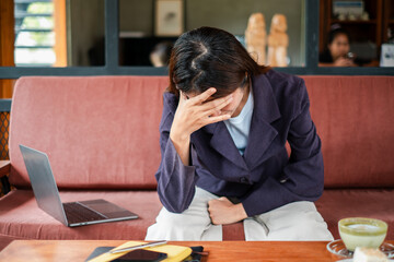 A woman in a blazer appears stressed while sitting on a couch with a laptop and coffee in a cozy...
