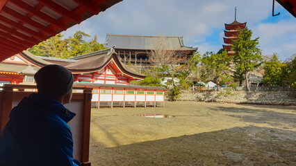 Lone visitor in a beanie, framed by a red gallery, looks out at Miyajima's Itsukushima Shrine and...