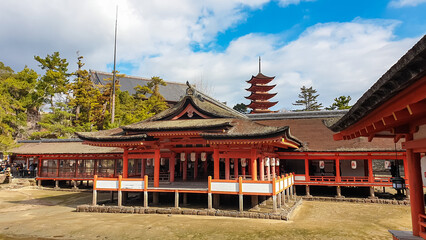 Itsukushima Shrine's vibrant orange wooden structures stand with Gojunoto Pagoda in the background, showcasing traditional Japanese architecture under a blue sky with white clouds in Miyajima, Japan
