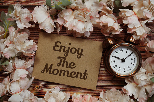 Naklejki Enjoy The Moment text with alarm clock and flowers decoration on wooden background