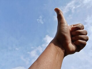 A man's hand shows a thumbs up gesture meaning OK with a clear sky in the background.