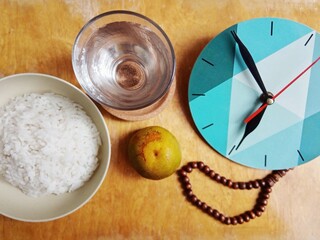 A glass of water, rice, and orange next to prayer beads and a clock on the dining table. The...