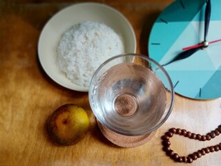 A glass of water, rice, and orange next to prayer beads and a clock on the dining table. The concept of breaking the fast. Flat lay view angle.