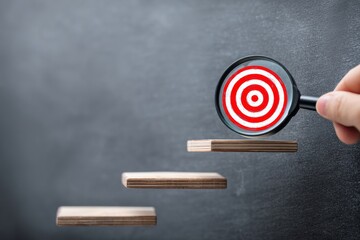 Hand using magnifying glass focusing on red target over wooden steps on blackboard background, representing goal setting and achievement