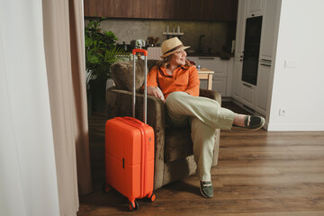 Smiling woman sits comfortably in a chair, excited for her summer vacation with an orange suitcase...