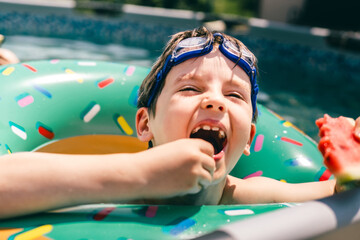 Happy boy in float eats watermelon and laughs in summer pool, enjoying sweet juicy fruit fun