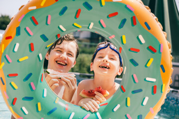 Two happy boys eating watermelon through colorful donut float on a hot summer pool day