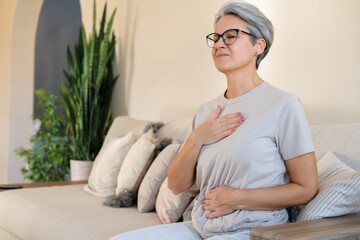 Senior woman feeling chest discomfort while sitting on sofa in cozy home environment