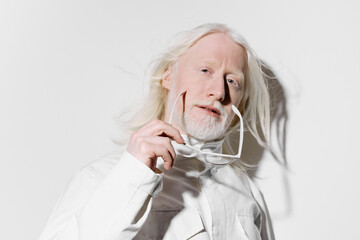Elderly man with long white hair and beard, holding glasses, against a plain white background,...
