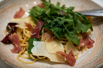 A pasta dish topped with arugula, prosciutto, and shaved Parmesan cheese