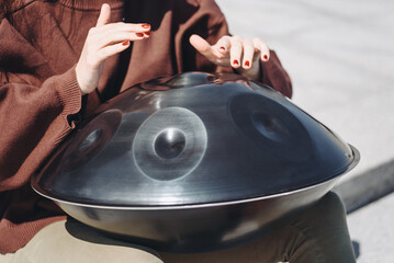 young caucasian woman playing black handpan with hands, ethnic metal drum instrument, sits on stone...