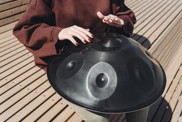 young caucasian woman playing black handpan with hands, ethnic metal drum instrument, sits on wooden bench of enbankment, beautiful relaxing music for meditation, close-up view of hands