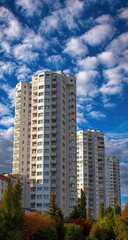 Fototapeta premium Tall, light-colored apartment buildings rise against a vibrant blue sky dotted with fluffy white clouds. A backdrop of trees and other buildings is visible at the base