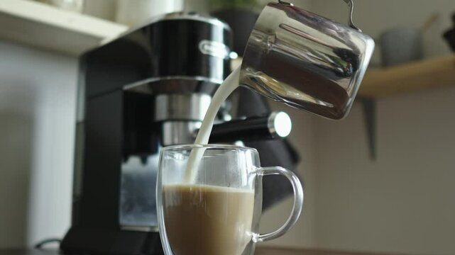 Milk pours from a pinscher into a glass cup of coffee. Cappuccino preparation. There is a coffee machine in the background