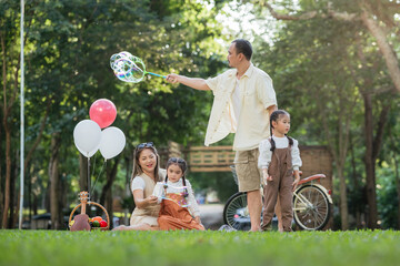 A family of four is sitting on the grass in a park. A man is holding a bubble wand and blowing...