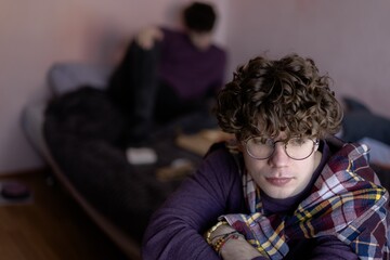 Obraz premium Young man with curly hair gazes thoughtfully while friend relaxes on bed in cozy, softly lit room during quiet afternoon