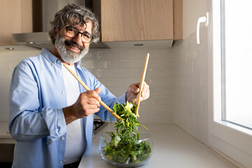 Happy mature man mixing bowl of fresh salad looking at camera smiling. Copy space