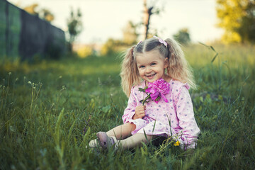cheerful 3-year-old girl with pigtails in a pink dress with a peony in a village meadow