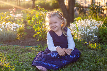 cheerful 3-year-old girl in a blue dress walks near the flowering flower bed in the backyard