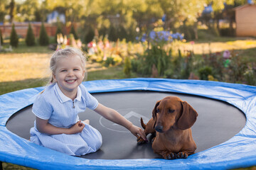 a three year old smiling girl playing with dachshund dog on trampoline in green sunny summer park