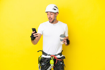 Young rock- climber man isolated on yellow background holding coffee to take away and a mobile