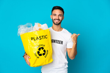 Young caucasian man holding a bag full of plastic bottles to recycle isolated on blue background...