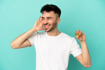 Young man holding a Bitcoin isolated on blue background listening to something by putting hand on...