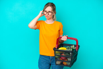 Blonde English young girl holding a shopping basket full of food isolated on blue background...