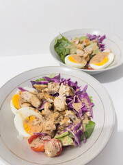 Healthy salad with boiled eggs, chicken pieces, cherry tomatoes, purple cabbage, lettuce, and sesame dressing. Fresh and nutritious meal in white bowls on a clean white background.