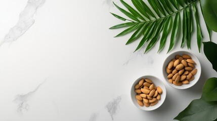 Flat lay of two small white bowls filled with almonds on a white marble background. on the right side of the image, there are two green palm leaves, one on the left side and one in the center.
