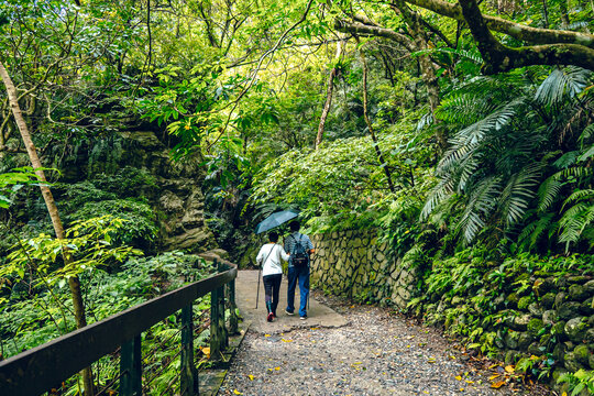 An older couple walks along a metal barrier walkway between mossy rocks. She carries a grey backpack and a beige bucket hat, surrounded by lush green trees in Taroko National Park, Taiwan.