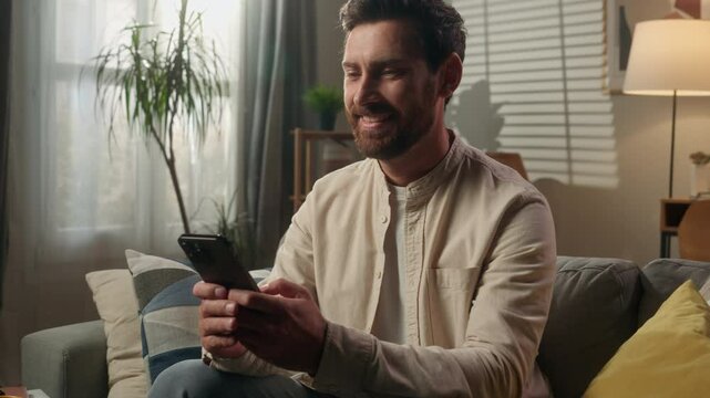Smiling man gadget addict businessman Caucasian guy homeowner relax using smartphone on couch living room happy male browsing mobile phone online shopping social media laughing enjoy message positive - Powered by Adobe