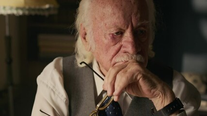 Medium close up shot of pensive senior Caucasian man wearing classic vest and holding eyeglasses sitting in retro home and contemplating about life - Powered by Adobe