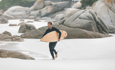 Adult male wearing black wetsuit walking across white sand carrying wooden surfboard with leash