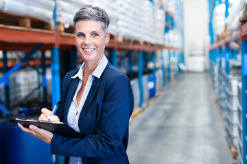 Middle-aged woman in blazer standing in warehouse writing on clipboard near racks, copy space