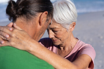 Senior woman and middle-aged man embracing and touching foreheads on sandy beach by ocean waves
