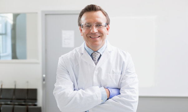 Male doctor standing arms crossed in clinical room wearing lab coat safety glasses and blue gloves