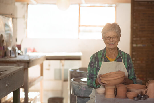 Senior woman wearing apron holding clay bowl at pottery wheel smiling in studio, copy space