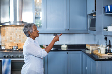 Senior Asian woman pointing at upper cabinet in modern kitchen with stainless steel kettle
