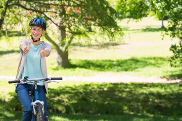 Woman wearing cycling helmet and cardigan straddling mountain bike in park giving thumbs up