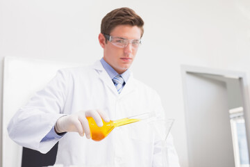 Male scientist in white coat pouring orange liquid through funnel into glass container on lab bench