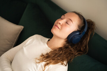 Top view young calm woman listening music in wireless headphones close eyes sitting on sofa at home enjoy the sound wear casual clothes rest spend free spare time in living room indoors, portrait