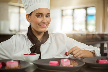 Female chef wearing uniform arranging raspberries on pink mousse on black plates at prep counter
