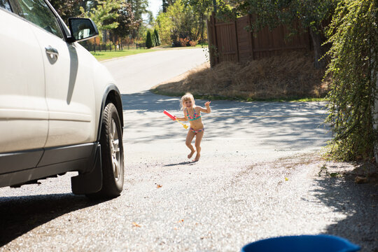 Female child wearing swimsuit crossing driveway holding water blaster near blue bucket and SUV