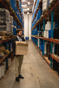 Man in business attire holding cardboard box, inspecting barrels on metal racks in warehouse aisle