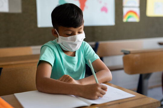 Asian boy wearing face mask writing in notebook at wooden desk in classroom with green pencil