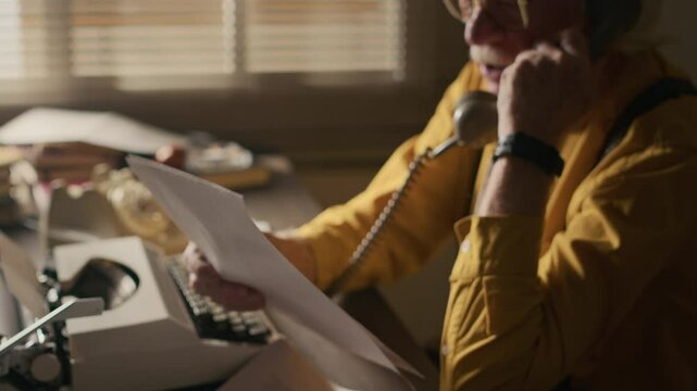 Side view shot of senior Caucasian male author holding paper script discussing deadline of his new book with publisher talking on retro rotary phone in his home study in front of vintage typewriter