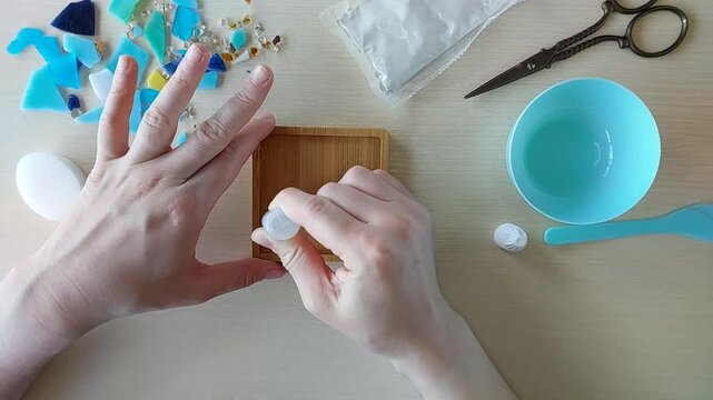 Spreading glue across wooden square base in preparation for placing colorful mosaic fragments for DIY gypsum coaster crafting project at home